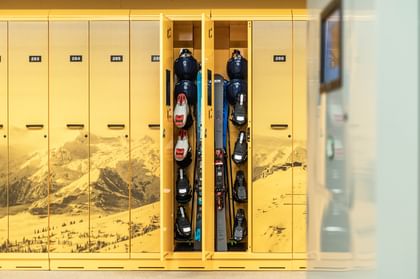 Yellow ski depot lockers with a mountain graphic. Two open lockers contain stored skis and ski boots.