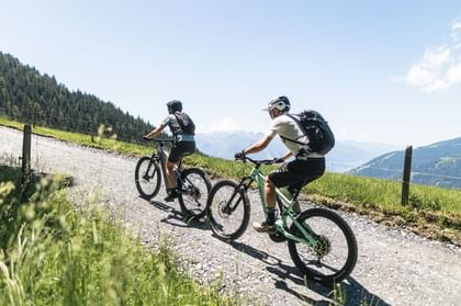 Two people wearing helmets and backpacks ride mountain bikes one behind the other along an uphill gravel trail. A fence runs alongside the path, and green hills and mountains can be seen in the background under a clear blue sky.