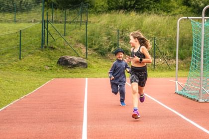 Auf dem Bild laufen zwei Kinder auf einer Laufbahn – ein Mädchen vorne in Sportkleidung, ein kleinerer Junge mit Kappe und Trainingsanzug hinter ihr. Beide schauen sich lächelnd an und bewegen sich auf ein Fußballtor am Rand der Bahn zu.