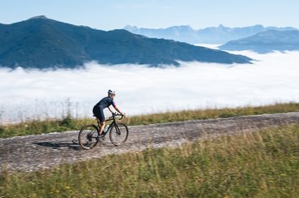 Eine Person mit weißem Helm und dunkler Radbekleidung fährt auf einem Schotterweg durch eine grüne Berglandschaft. Unterhalb der Berge liegt eine dichte Nebelschicht, darüber erstreckt sich ein klarer Himmel.