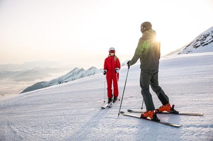 Das Bild zeigt zwei Skifahrer auf einer verschneiten Piste, umgeben von Bergen. Eine Person trägt eine rote Skiausrüstung, die andere eine grüne. Sie unterhalten sich bei Sonnenaufgang oder Sonnenuntergang in einer idyllischen Winterlandschaft.