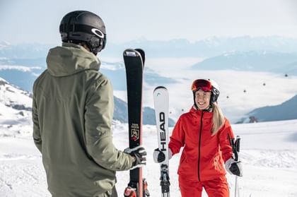 Eine Frau in roter Skiausrüstung und ein Mann in grüner Skiausrüstung unterhalten sich auf der Piste, beide halten ihre Skier.