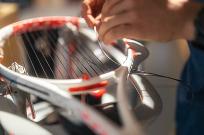 Das Bild zeigt eine Nahaufnahme eines Tennisschlägers während des Besaitungsvorgangs. Eine Hand führt gerade eine Saite durch den Rahmen, während der Schläger in einer Besaitungsmaschine eingespannt ist.