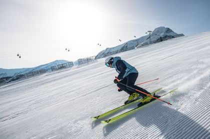 Ein Skifahrer in blauer Skiausrüstung fährt eine frisch präparierte Piste hinunter. Im Hintergrund sind schneebedeckte Berge, ein Seilbahnträger und Gondeln zu sehen.