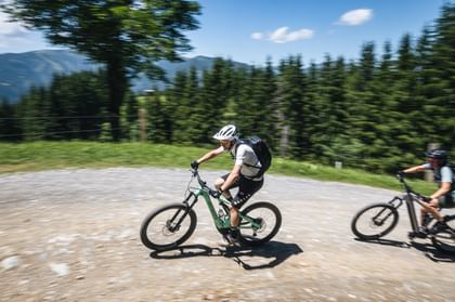 Zwei Personen mit Helmen und Rucksäcken fahren mit Mountainbikes durch eine Kurve auf einem breiten Schotterweg. Im Hintergrund sind grüne Bäume, ein Zaun und Berge unter einem klaren blauen Himmel zu sehen, während die Bewegung leicht verschwommen wirkt.