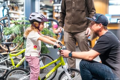 Das Bild zeigt ein kleines Mädchen in der Bründl Sports Bikeworld, das auf einem Kinderfahrrad steht, während ein Mitarbeiter kniend die Einstellungen überprüft und ein Erwachsener zuschaut.