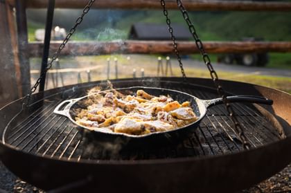 Fresh Kaiserschmarrn dusted with powdered sugar is prepared in a cast-iron pan over an open fire. A green alpine meadow is visible in the background.