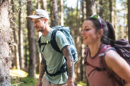 Two people with backpacks are walking through a forest with tall, slender trees. The man wears a light-colored cap and a green T-shirt, the woman a sleeveless top; sunlight filters through the trees onto the forest floor.