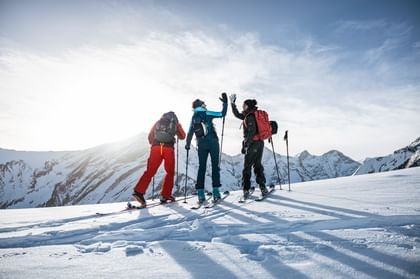 Drei Personen in Skitouren-Ausrüstung stehen auf einem verschneiten Bergrücken, während zwei von ihnen sich fröhlich mit einem High-Five abklatschen, umgeben von einer beeindruckenden Berglandschaft.