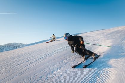 Ein Skifahrer fährt in enger Kurvenlage über eine präparierte Skipiste und wirbelt Schnee auf. Ein weiterer Skifahrer ist im Hintergrund zu sehen.