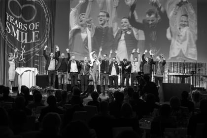 Eine Gruppe von Personen steht vor Publikum auf einer Bühne und hebt jubelnd die Arme. Im Hintergrund wird die Gruppe auf einer großen Leinwand gezeigt, seitlich ist der Schriftzug „60 Years of Smile“ zu sehen.