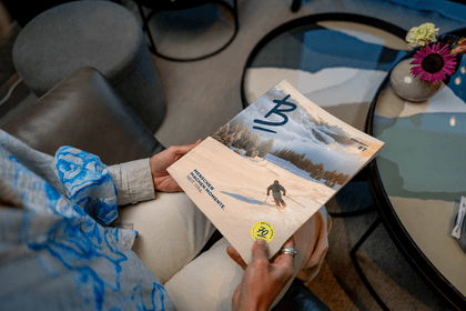 A seated person holds the Bründl Sports magazine featuring a skier on a snowy slope. The cover includes the text “Menschen machen Momente. Seit 1956.”, “Bründl Sports #7” and a yellow 70-year emblem.