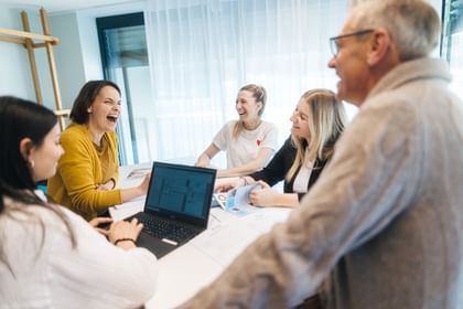 Five people are sitting around a table in a bright room, laughing together. A laptop and several documents are on the table, being looked at by the group.