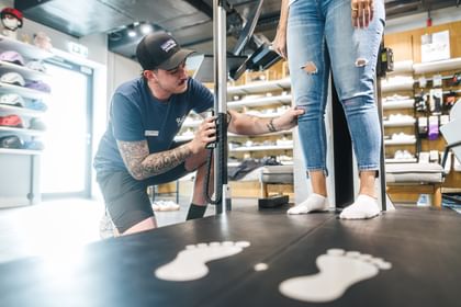 A Bründl Sports employee measures a customer’s leg position during a bike fitting. The customer stands in socks on a measuring platform, with shoes and sports products visible in the shop background; a Patagonia logo can be seen on the employee’s cap.