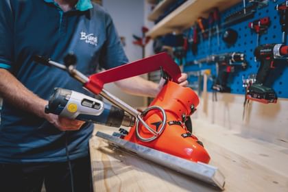 Eine Person erwärmt in einer Werkstatt eine leuchtend orange Skischuhschale mit einem Heißluftgerät, das auf die Außenseite des Schuhs gerichtet ist. Der Skischuh ist auf einer Metallvorrichtung fixiert, im Hintergrund sind Werkzeuge an einer blauen Wand zu sehen.