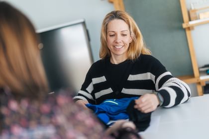 A woman holds a blue piece of clothing and shows it to another person at the table. She is smiling, with a screen and a board visible in the background.
