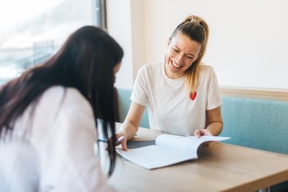 Two women are sitting at a table in a bright room. One woman is smiling and pointing at an open document, while the other is looking down and writing.