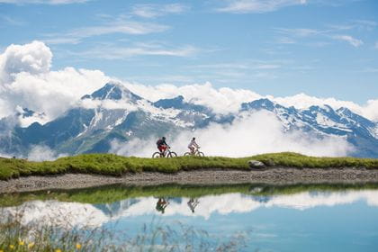 two biker are riding alongside a mountain lake, wherein the mountains are mirrored.
