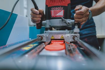 Eine Person bedient in einer Werkstatt eine Fräsmaschine, durch die ein leuchtend rotes Kunststoffteil geführt wird. An den Seiten sind Metallschienen, Warnsymbole und Kunststoffspäne zu sehen.