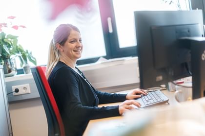 A woman sits on a red office chair at a desk typing on a keyboard in front of a monitor. She is smiling slightly, with a window and plants visible in the background.
