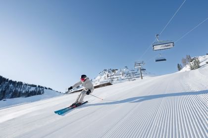 Ein Skifahrer fährt in einer Kurve über eine frisch präparierte Skipiste. Im Hintergrund sind ein Sessellift, verschneite Berghänge und ein klarer blauer Himmel zu sehen.