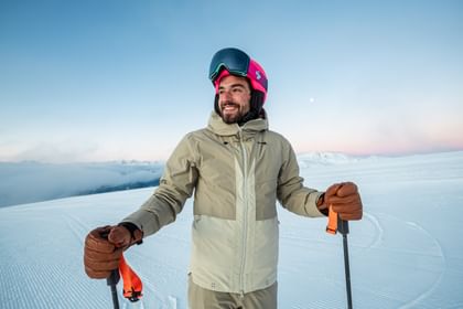 Ein lächelnder Skifahrer mit pinkem Helm und Skibrille steht auf einer verschneiten Piste bei Sonnenaufgang, mit Blick auf die umliegenden Berge.
