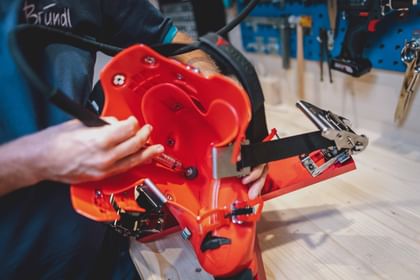 Eine Person bearbeitet in einer Werkstatt eine leuchtend rote Skischuhschale, die auf einer Werkbank fixiert ist. Im Hintergrund sind verschiedene Werkzeuge und eine blaue Werkzeugwand zu sehen.