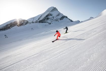 Zwei Skifahrer fahren eine präparierte Piste hinunter, umgeben von einer sonnigen Berglandschaft.