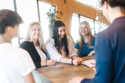 Several people stand at a table looking at a document together. One person points at it while the others listen and smile.