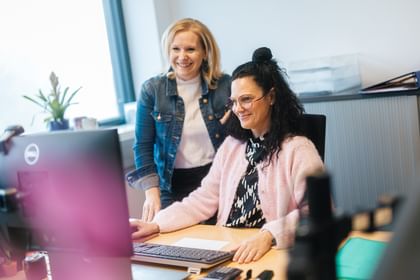 One woman sits at a computer using a keyboard and mouse, while another woman stands behind her looking at the screen. Both are smiling, and papers and office supplies are on the desk.