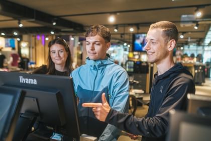 Drei Personen stehen in einem Sportgeschäft vor einem Bildschirm. Ein Mitarbeiter zeigt mit der Hand auf den Monitor, während die beiden anderen Personen aufmerksam darauf schauen. Im Hintergrund sind beleuchtete Verkaufsflächen und Sportbekleidung zu sehen.