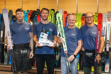 Vier Mitarbeiter von Bründl Sports stehen vor einer Skiwand und präsentieren verschiedene Ski sowie einen Skischuh.
