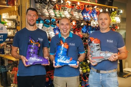Drei Mitarbeiter von Bründl Sports stehen vor einer Wand mit Skischuhen und präsentieren Modelle von Tecnica, Lange und Rossignol.