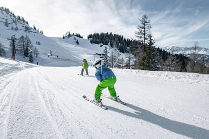 Two children ski down a snowy slope, one behind the other. The child in front wears a blue jacket and green pants; trees, a ski lift and mountains are visible in the background.