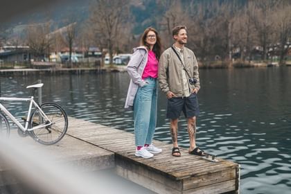 Eine Frau mit Brille und ein Mann stehen nebeneinander auf einem Holzsteg am Wasser und schauen in die Ferne. Links ist ein Fahrrad zu sehen, im Hintergrund ein ruhiger See, Bäume und einige Gebäude.