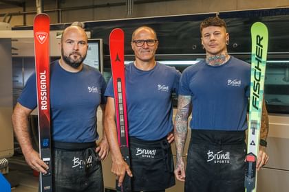 Drei Mitarbeiter von Bründl Sports stehen in der Skiwerkstatt und halten Rennski von Rossignol, Atomic und Fischer.