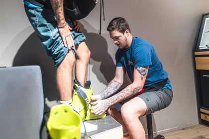 Bootfitting A Bründl Sports employee fits a yellow ski boot for a customer. The employee kneels down to ensure the perfect fit while the customer stands on a bench.