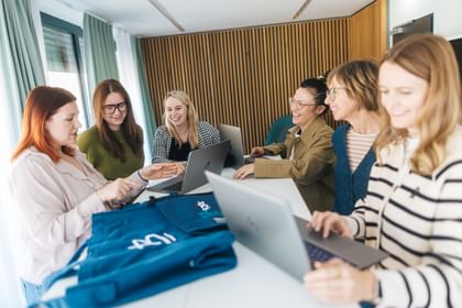 Several women are standing and sitting at a table with open laptops, looking at blue clothing items. One woman points at a detail while the others smile and look at the clothing.