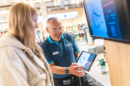 Bootfitting A Bründl Sports employee advises a customer in the shop, showing a digital ski boot analysis on a tablet. Sports equipment and ski gear are visible in the background.