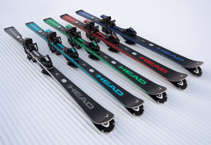 Fünf Paar schwarze HEAD Ski mit Bindungen liegen nebeneinander auf präpariertem Schnee. Die Ski tragen große HEAD-Schriftzüge und Details in Grau, Türkis, Grün, Rot und Blau.