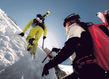 Skitourengeher beim Aufstieg im Hochgebirge mit Dynafit-Ausrüstung Zwei Skitourengeher steigen mit Eispickeln und aufgeschnallten Skiern eine verschneite, steile Passage hinauf. Beide tragen Dynafit-Bekleidung und Helme.