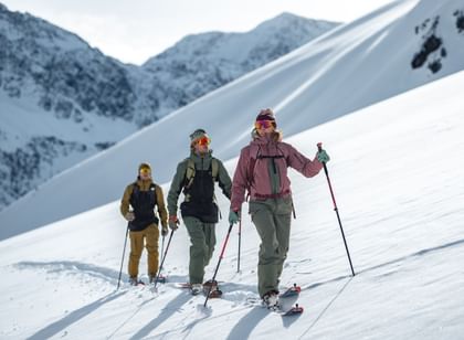 Das Bild zeigt drei Skifahrer in Winterkleidung, die mit Skistöcken und Tourenski mit Fellen einen verschneiten Berg hinaufwandern. Im Hintergrund ist eine beeindruckende Berglandschaft zu sehen.