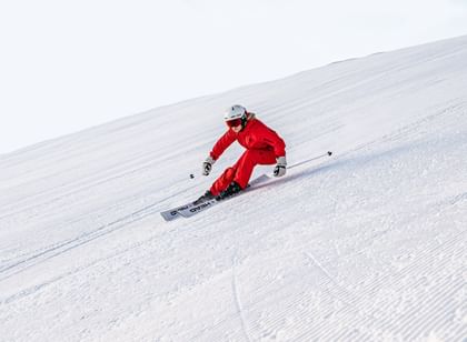 A female skier in a red Scott ski suit carves turns through the snow on a white Head ski. She is also wearing a white POC ski helmet and black Oakley ski goggles.
