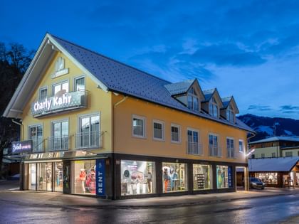 Das Bild zeigt ein Gebäude mit dem "Bründl Sports" Shop im Erdgeschoss und einem Schild für "Charly Kahr" im oberen Bereich, umgeben von anderen Gebäuden in einer winterlichen Umgebung.