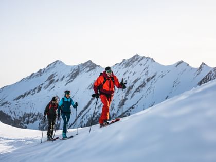 Drei Personen in Skitouren-Ausrüstung steigen eine verschneite Bergflanke hinauf, während im Hintergrund majestätische, schneebedeckte Berge zu sehen sind.<br/><br/>