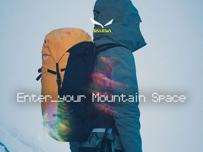 A person with their hood up wears a yellow-and-black backpack and climbing equipment attached to their harness against a snowy background. The image features the Salewa logo and the text “Enter your Mountain Space”.