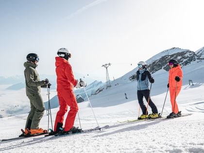 Das Bild zeigt vier Skifahrer, zwei Männer und zwei Frauen, die auf einer Skipiste stehen und sich unterhalten. Sie tragen bunte Skiausrüstungen (grün, rot, orange, blau), umgeben von einer verschneiten Berglandschaft.