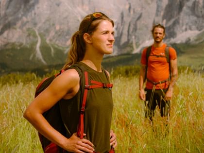 Eine Frau mit rotem Rucksack steht selbstbewusst in einer Almwiese vor beeindruckender Bergkulisse, während ein Mann in orangem Shirt im Hintergrund steht. Beide tragen funktionale Wanderkleidung und genießen die Aussicht.