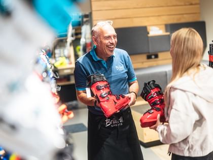 Ein Bründl Sports Mitarbeiter berät eine Kundin bei der Auswahl von Skischuhen. Beide halten rote Skischuhe in der Hand und lachen, während sie sich im Shop umsehen.