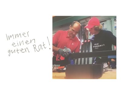 Zwei Männer arbeiten gemeinsam an einem Ski in einer Werkstatt, im Hintergrund stehen weitere Ski. Daneben steht handschriftlich „Immer einen guten Rat!“.